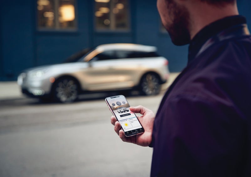 A man is holding his phone looking at the Lincoln Way™ App and we see his 2022 Lincoln Corsair SUV in the background against a blue brick wall | Bob Maxey Lincoln in Detroit MI