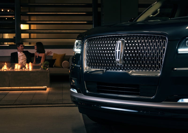 The signature Lincoln grille on the 2022 Lincoln Navigator SUV commands attention as a couple relaxes by a fire beyond | Bob Maxey Lincoln in Detroit MI