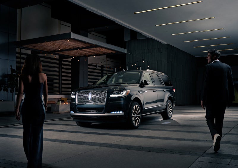 A man and woman in formal wear are approaching a 2022 Lincoln Navigator SUV at night as it lights with the Lincoln Embrace | Bob Maxey Lincoln in Detroit MI
