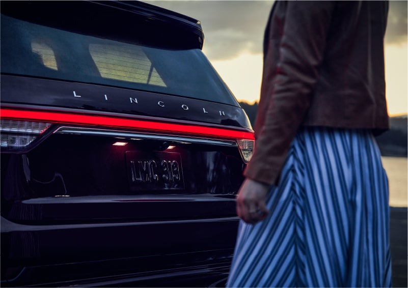 A person is shown near the rear of a 2023 Lincoln Aviator® SUV as the Lincoln Embrace illuminates the rear lights | Bob Maxey Lincoln in Detroit MI